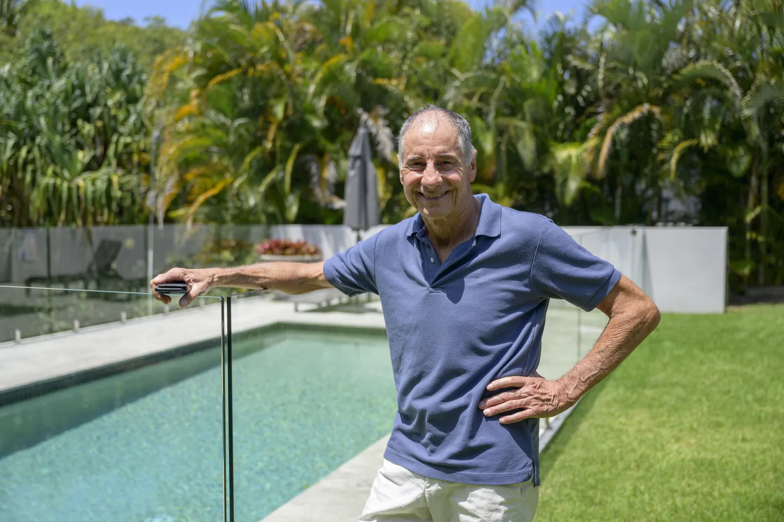 A smiling retired man holding his phone beside his fenced backyard pool